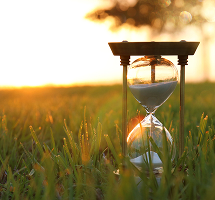 Close up of an hourglass sitting in a field with the sun setting behind it.