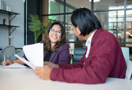 A smiling woman facing the camera sits with a man holding papers.