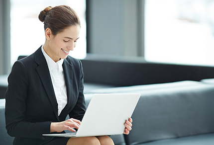 A smiling young professional sitting in a lobby looking at an open laptop.