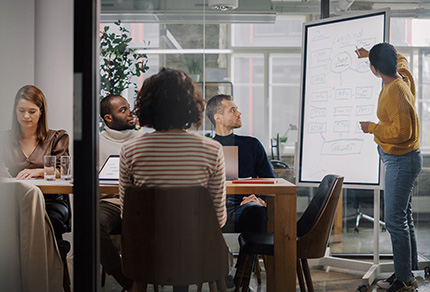 A group of business people sitting around a meeting table while a woman writes on a white board.
