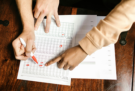 People highlighting and pointing at a spreadsheet sitting on a wooden table.
