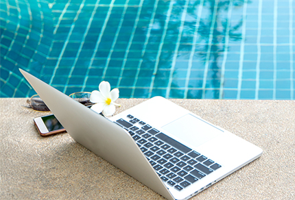 A laptop, iPhone, sunglasses and a flower sitting next to an outdoor swimming pool.