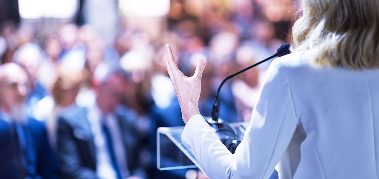 A presenter speaking at a podium in front of a large audience.