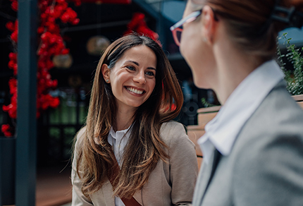 A smiling professional woman talking to another professional in an outdoor setting.